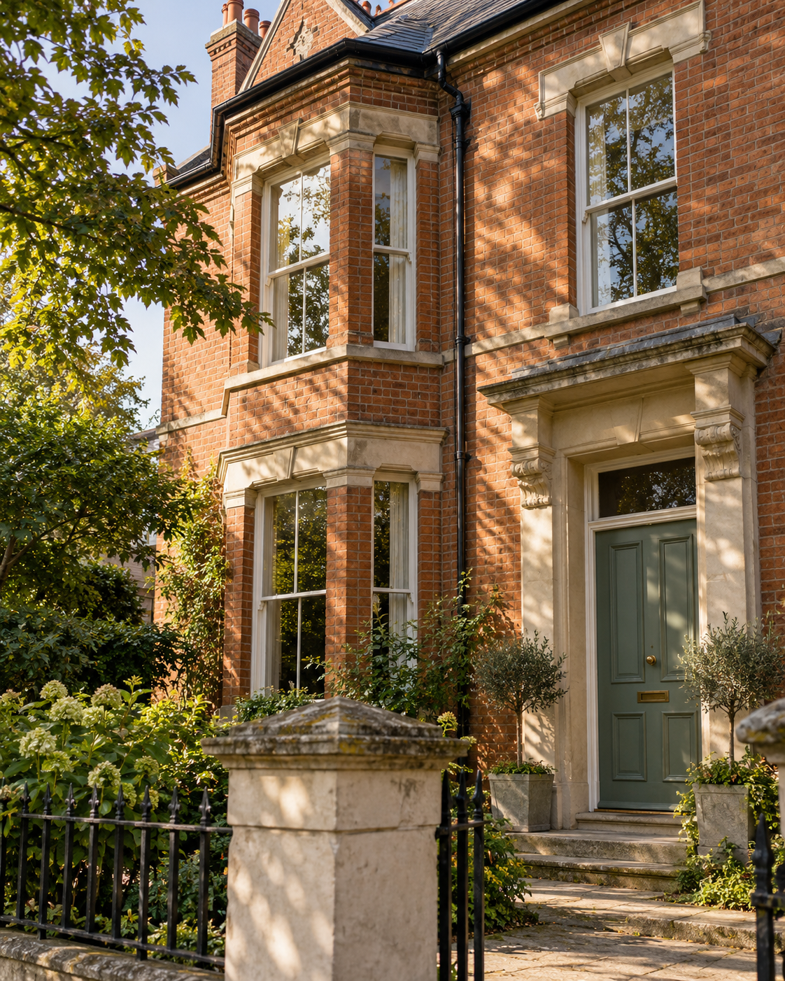 Sunlight and tree shadows across a Victorian brick home with sash windows and a sage-green door